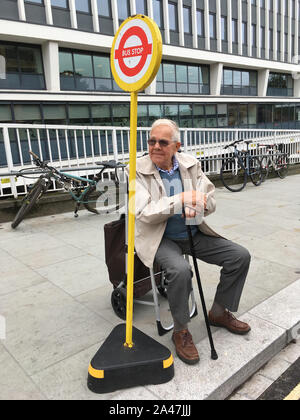 Ältere Männer/älterer Mann wartet geduldig auf seinem tragbaren push Gestell für seinen Bus an seine temporäre Bushaltestelle auf dem Bürgersteig zu gelangen. Twickenham. UK. (Stockmo) Stockfoto