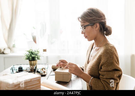 Junge Frau in casualwear sitzt von Tisch und verbindlichen Knoten auf der Oberseite der Verpackung Stockfoto