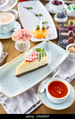 Stück leckeren Käsekuchen mit Erdbeeren und Minze auf weiße Platte. Frühstück im Café, Kaffee am Morgen. Cappuccino und viele Desserts Stockfoto