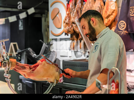 Madrid, Spanien - 4. Juni 2017: Metzger schneiden eine ganze Bein der serrano Iberico Schinken auf einem Ständer für Kunden auf einem Markt, Schinken hängen im Hintergrund Stockfoto