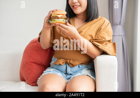 Hungrig übergewichtige Frau und lächelnd Hamburger und sitzen im Wohnzimmer, sie sehr glücklich und genießen Sie Fast Food zu essen. Konzept der binge eati Stockfoto