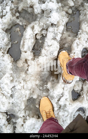 Person im gelben Stiefel versucht, einen Schritt durch Schneematsch auf einer verschneiten Straße und als Hindernis für die Passage von Fußgängern, gedruckten Fußspuren und Wat Stockfoto