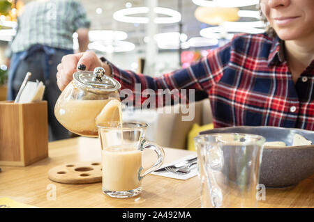 Mittagessen im Restaurant. Eine hübsche Frau lächelt im Vorgriff auf eine indische masala trinken, die Sie aus einer Teekanne aus Glas in eine Schale gießt, neben ihr Stockfoto