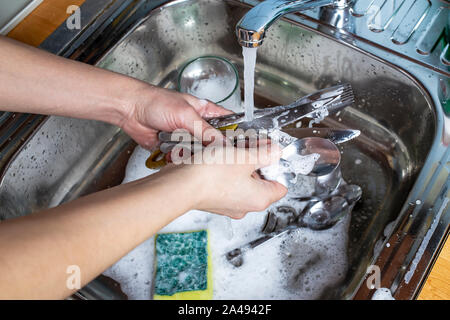 Menschliche Hand waschen Löffel, Gabeln, Messer mit einem Schwamm und Spülmittel unter fließendem Wasser in der Spüle, Geschirr und Glas. Stockfoto