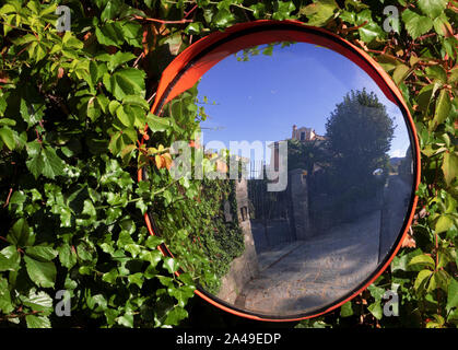 Konvexe Spiegel hängen von einem Zaun mit Blättern bedeckt, spiegelt die Landschaft in einer engen Straße in Italien Stockfoto