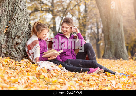 Freizeitaktivitäten im Freien. Schwestern sitzen in der Nähe von Tree unter Decke im Herbst park Lesung buchen Sie lächelt glücklich Stockfoto