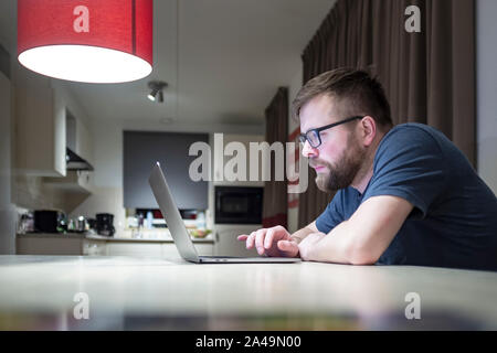 Konzentriert, hübscher junger Blogger mit Bart und Brille, zeichnet die Entwicklung von seiner Seite, zu Hause sitzen am Tisch und schaut auf einen Laptop in einer Co Stockfoto