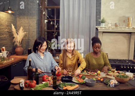 Hohen winkel Portrait von drei jungen Frauen kochen Abendessen für House party beim Stehen in der Reihe an den Tisch und Schneiden von Gemüse, kopieren Raum Stockfoto