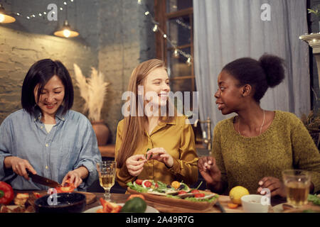 Portrait von drei jungen Frauen kochen Abendessen für House party, während in der Zeile in der Tabelle steht und munter plaudernd Stockfoto