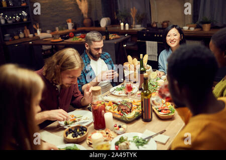 Multi-ethnischen Gruppe von Freunden genießen Sie köstliche Abendessen zusammen am Tisch sitzen in schwach beleuchteten Zimmer, auf lächelnden Asiatin Fokus Stockfoto