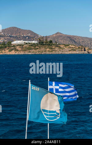 Elounda, Kreta, Griechenland. 2019. Die Blaue Flagge für einen ausgezeichneten Strandflug mit der Nationalflagge Griechenlands in Elounda, Kreta. Stockfoto
