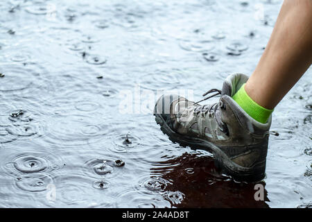 Frau in touristische wasserdichte Wanderschuhe Wandern am Wasser in Pfützen im Regen. Close-up. Stockfoto