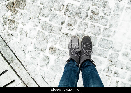 Die Beine der Frauen in der stilvollen Wildleder Stiefel stehen auf der schneebedeckten Fahrbahn aus Granit Pflastersteine, neben ist ein Abflusssystem. Ansicht von oben. Stockfoto