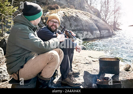 Zwei glückliche Reisende sitzen auf der felsigen Ufer des Sees, Chatten, lebhaften und trinken Bier durch das Feuer, auf denen das Essen in einem Topf zubereitet wird. Clo Stockfoto