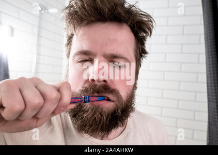 Eine verschlafene Mann mit geschlossenen Augen, zerzausten Haaren und einem dicken Bart ist seine Zähne putzen mit der Zahnbürste, am frühen Morgen, im Bad. Stockfoto