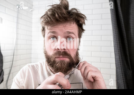 Der Mensch ist in seinem shaggy in den Spiegel schauen, überrascht, nahm die Schere in der einen Hand und in einem anderen Teil der Bart und mit einer ernsthaften Absicht Ri ist Stockfoto