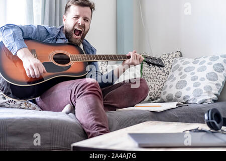 Eine junge, gut aussehende männliche Komponisten spielt eine akustische Gitarre und lauthals singt ein Lied seiner eigenen Komposition, sitzen auf einem Sofa, in ein gemütliches Heim envi Stockfoto