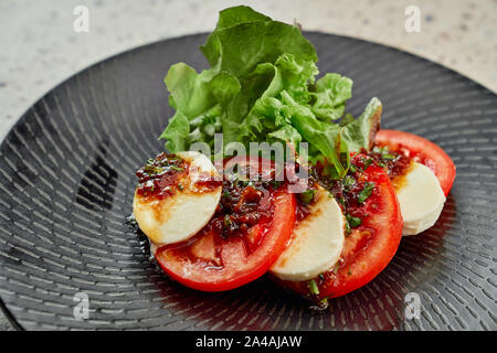 Lecker Caprese Salat aus Schichten von Tomaten, Mozzarella und grünem Salat, ausgegossen mit Soße, auf schwarzem Teller serviert. Stockfoto