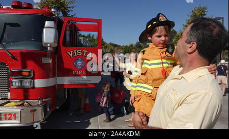 Ein Papa trägt seinen kleinen Jungen als Feuerwehrmann gekleidet, wie sie die Fahrzeuge im Fire Station open house erkunden Stockfoto