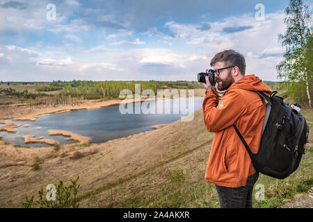 Ein bärtiger Mann - ein Tourist mit einem Rucksack steht auf einem Hügel, hält eine Kamera in den Händen und nimmt Bilder einer schönen Landschaft, auf einem autolöscher Stockfoto