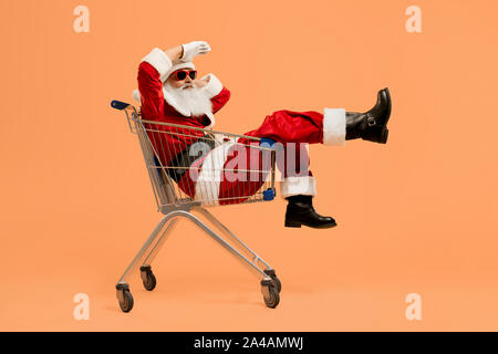 Echte reife Santa Claus mit grauen Bart in trendigen Sonnenbrille weg schauen, während im Warenkorb liegen aus dem Supermarkt und die Pause während der harten Arbeit. Weihnachten und das neue Jahr. Stockfoto