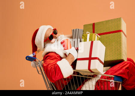 Authentische Santa Claus mit grauen Bart in trendigen Sonnenbrille im Warenkorb mit vielen farbigen Geschenkboxen und rotem Papier Cup. Auf orange studio Hintergrund isoliert. Stockfoto