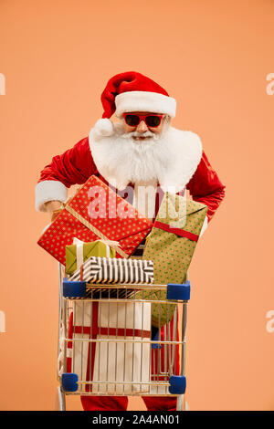 Glücklich lächelnde senior Santa Claus mit echten grauen Bart rot Sonnenbrille an Kamera suchen und gleichzeitig den Warenkorb mit bunten Geschenkboxen. Weihnachten Vater Geschenke geben, glückliche Kinder. Stockfoto