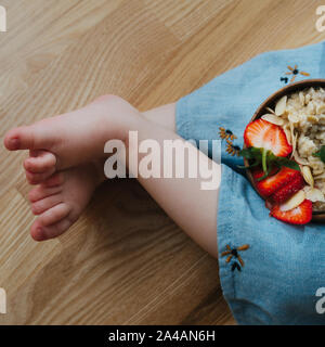 Nahaufnahme der Beine kleines Mädchen und ein Teller Müsli mit Bananen und Erdbeeren Stockfoto