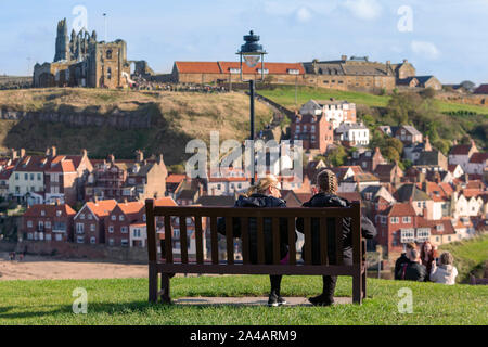 Zwei Frauen sitzen auf einer Holzbank mit Blick auf Menschen mit der 199 Schritte zu Whitby Abbey und die Kirche der Heiligen Maria Stockfoto