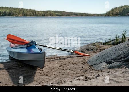 Kajaks mit Paddeln liegen auf dem sandigen Ufer neben großen Steinen, die vor dem Hintergrund von einem See und einem felsigen Ufer mit Bäumen, an einem sonnigen Sommertag. Acti Stockfoto