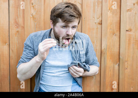 Bärtiger Mann reinigt seine Brille aus Staub und wird Sie mit einem T-Shirt abwischen. Auf dem Hintergrund einer alten hölzernen Wand isoliert. Close-up. Stockfoto