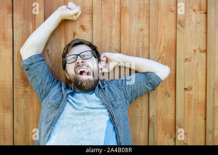 Eine entspannte bärtig, kaukasischer Mann lustig gähnt und streckt sich, im Freien im Sommer. Isolierte, auf dem Hintergrund der gemalten alte Holz- Oberfläche. Schließen Stockfoto