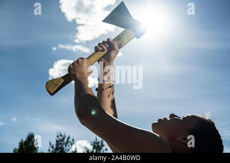 Hält eine Frau eine Axt in den Händen gegen den blauen Himmel, die Sonne und der Baumkronen. Konzept der Gleichstellung der Geschlechter und die moderne Frau. Close-up. Stockfoto