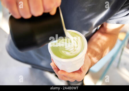Männliche Barista, vegan Matcha Cappuccino mit hafermilch mit latte Kunst im Café im Freien. Stockfoto