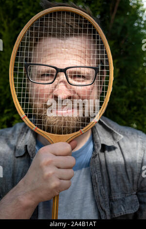 Eine freudige, lustige Mann Schutzbrille aufsetzen auf ein Badminton Schläger und lehnte sich gegen sein Gesicht für ein Witz. Close-up. Stockfoto