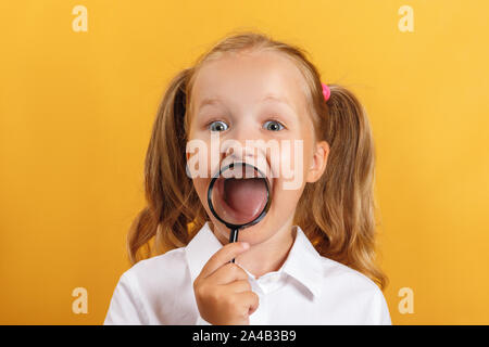 Fröhliche Schülerin kleines Mädchen zeigt Zähne und lächelt durch ein Vergrößerungsglas. Ein Kind auf einem gelben Hintergrund. Stockfoto