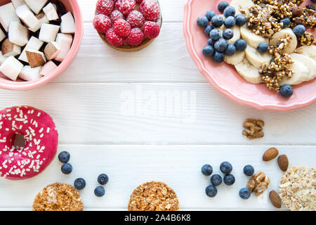 Gesunde knackige Kekse und Müsliriegel, Bananen und Kokos Stücke, rosa Donut und Himbeere tart auf weissem Holztisch. Pastellfarben, freier Speicherplatz für Stockfoto