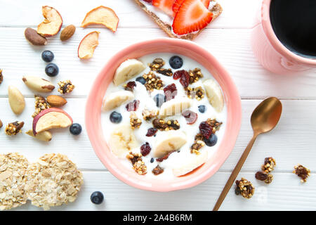 Schüssel mit gesunden Joghurt mit Früchten und Beeren, frische Bananen, getrocknete Früchte, serviert mit einer Tasse Kaffee schwarz auf weiß Holz- Hintergrund. Stockfoto