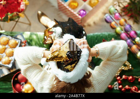 Urlaubszeit. Obere Ansicht für die moderne Hausfrau in glitzern gold Pailletten Weihnachtsmütze sitzen auf einem Sofa im Wohnzimmer zu Weihnachten. Stockfoto