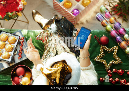 Urlaubszeit. Obere Ansicht der verärgerten stilvolle Frau in glitzern gold Pailletten Weihnachtsmütze sitzen auf einem Sofa im Wohnzimmer an Weihnachten anrufen ai c Stockfoto