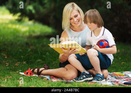 Liebevolle Mama liest ein Buch zu Ihrem besonderen Kind auf einem Park Rasen Stockfoto