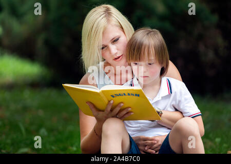 Junge Mutter Lesung ein englisches Buch zu Ihrem Kindergarten Sohn auf einem Park Rasen Stockfoto