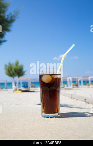 Glas Cuba Libre Cocktail mit Eis und Stroh auf dem weißen Sandstrand, Meerblick Hintergrund. Stockfoto