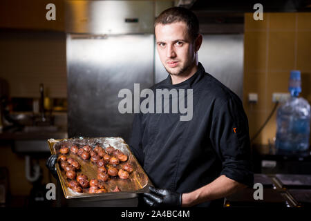 Portrait männlichen Chef mit gekochtem Essen in der Küche. Thema kochen. jungen kaukasischen Mann in schwarzer Uniform, Latex Handschuhe im Restaurant in der Küche Stockfoto