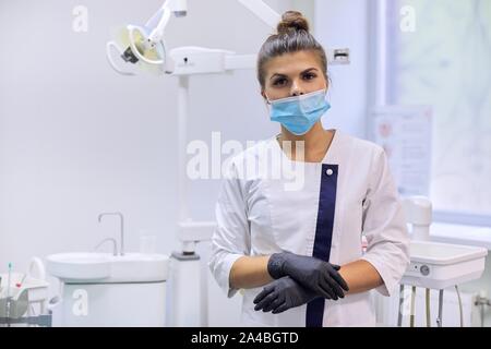 Portrait von zuversichtlich weiblichen Zahnarzt Arzt im weißen Kittel mit Maske in der Nähe von zahnmedizinischen Stuhl in der zahnmedizinischen Klinik Büro Stockfoto