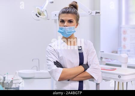 Portrait von zuversichtlich weiblichen Zahnarzt Arzt mit verschränkten Armen im weißen Mantel mit Maske in der Nähe von zahnmedizinischen Stuhl in der zahnmedizinischen Klinik Büro Stockfoto