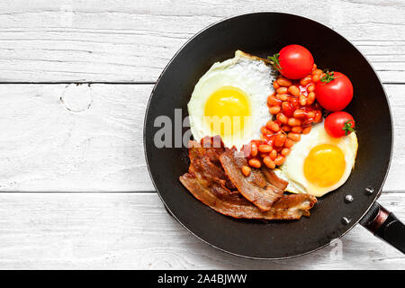 Englisches Frühstück in der Pfanne mit Spiegelei, Speck, Bohnen und Tomaten auf weißem Holz- Hintergrund. top View Stockfoto