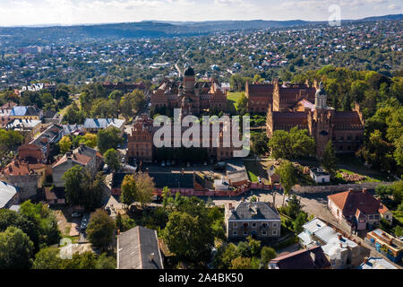 Bunte alte Gebäude der Czernowitzer Universität mit Türmen und Kuppeln zwischen grünen Bäumen und Häusern am Himmel Hintergrund in der Ukraine. Sonne scheint Stockfoto