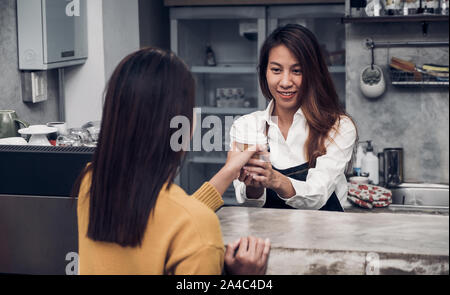 Junge asiatische Frau barista Schürze tragen Jean serviert Kaffee Tasse zu Kunden an Theke mit Smile Emotion zu gehen, Cafe Restaurant Service Konzept, Inhaber Stockfoto