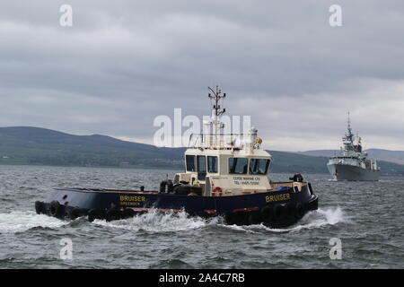 Bruiser, eine Damen Stan 1907 tug von Clyde Marine Services betrieben, Pässe Greenock mit hmcs St John's im Hintergrund. Stockfoto